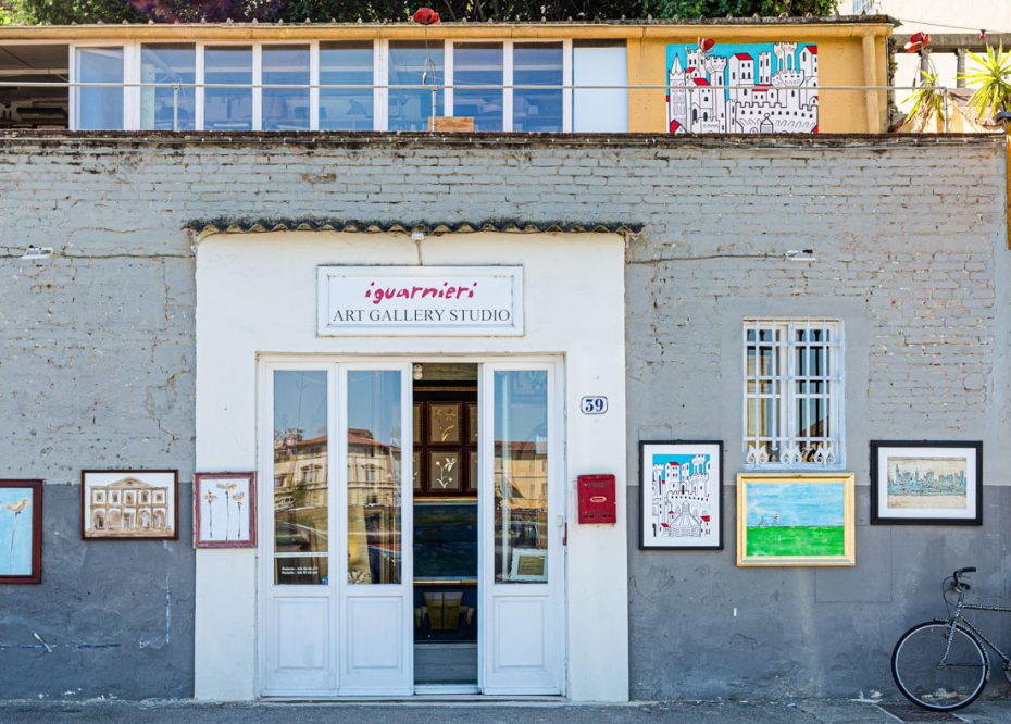 White double doors of an art gallery with framed paintings along the gray exterior wall; a bicycle rests nearby on the right.